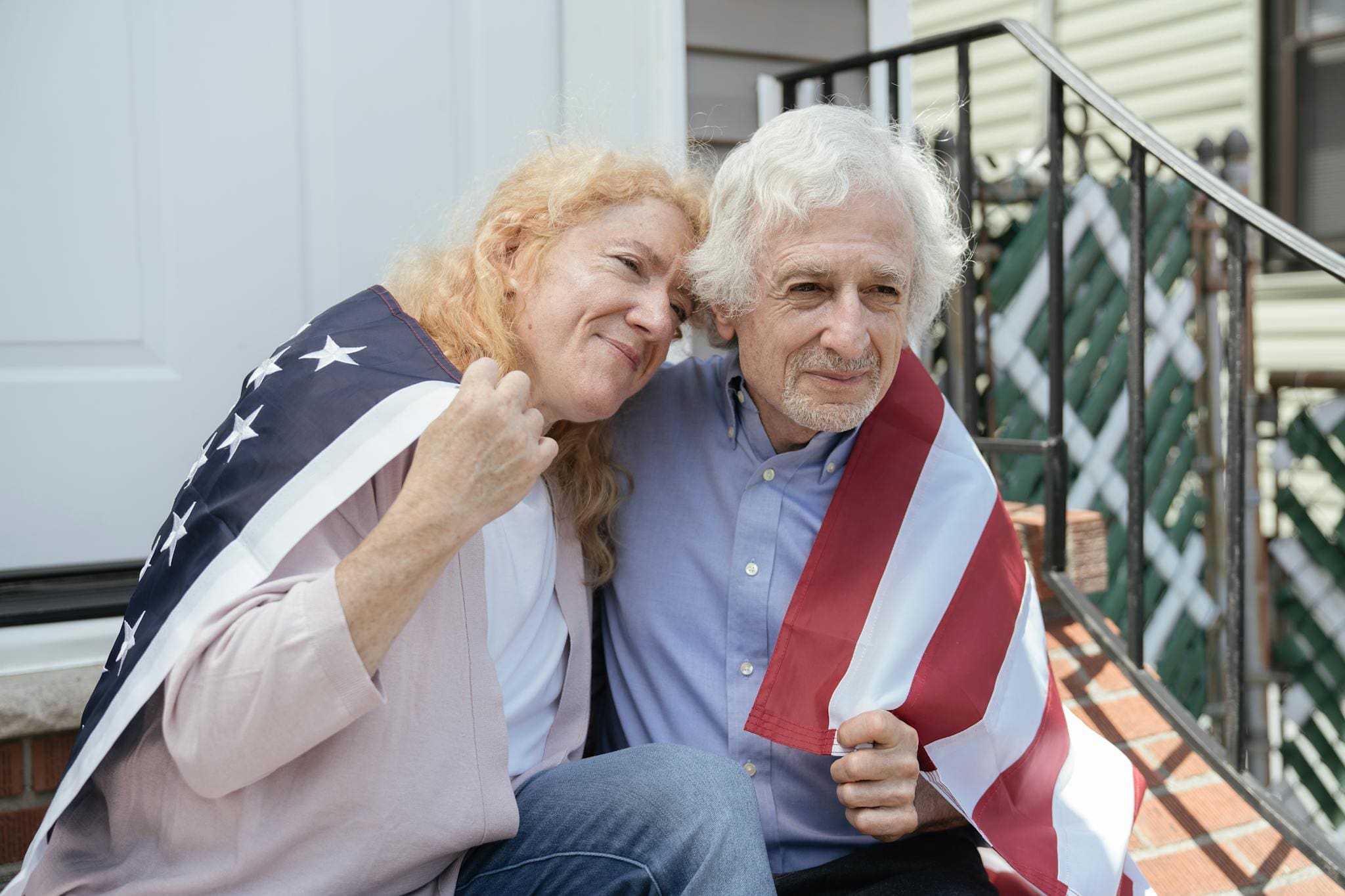 Joyful senior couple wrapped in American flag on porch steps celebrating Independence Day.